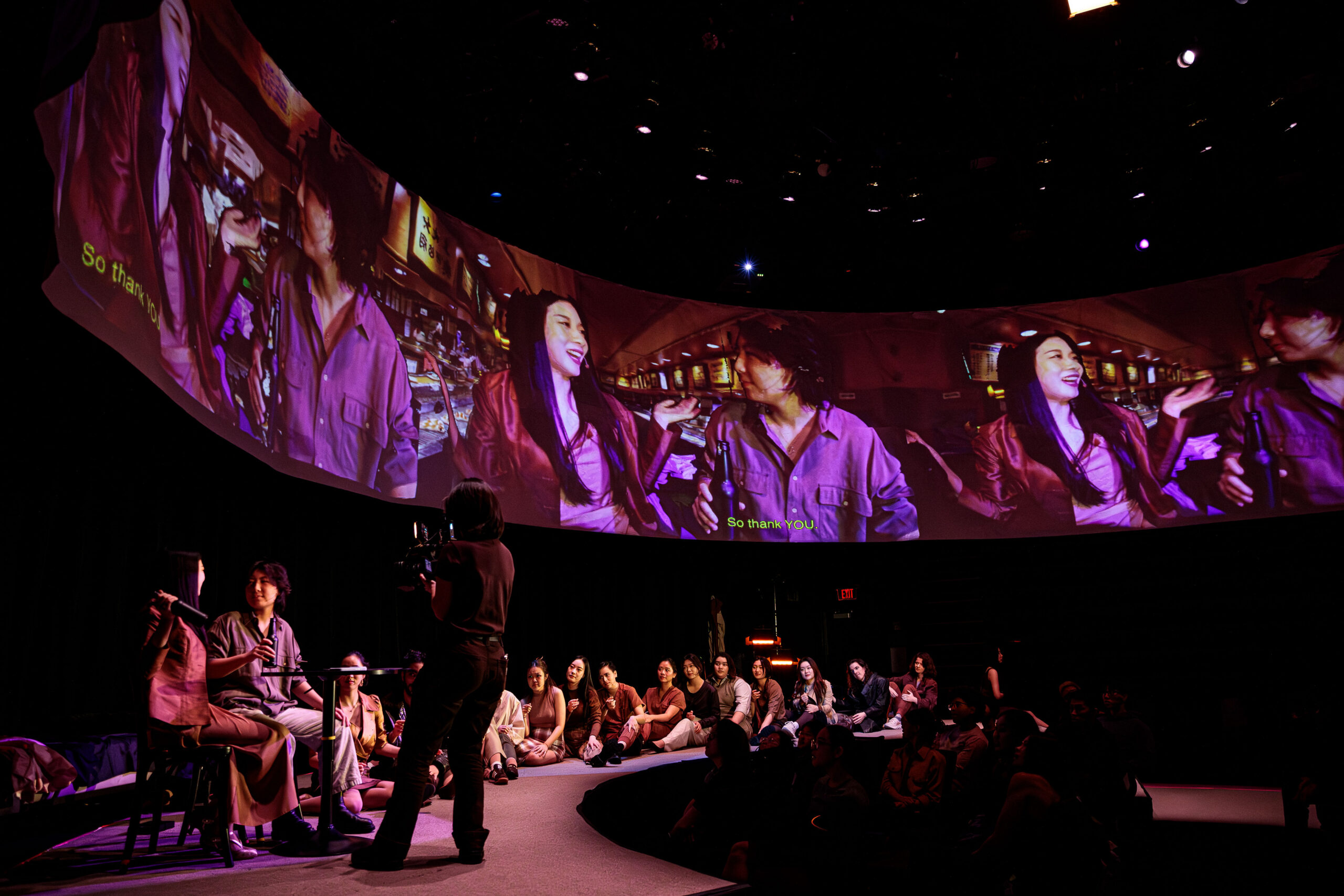Photo from a production of A Dream Like a Dream. Performers on a circular stage with a curved projection screen above them. 