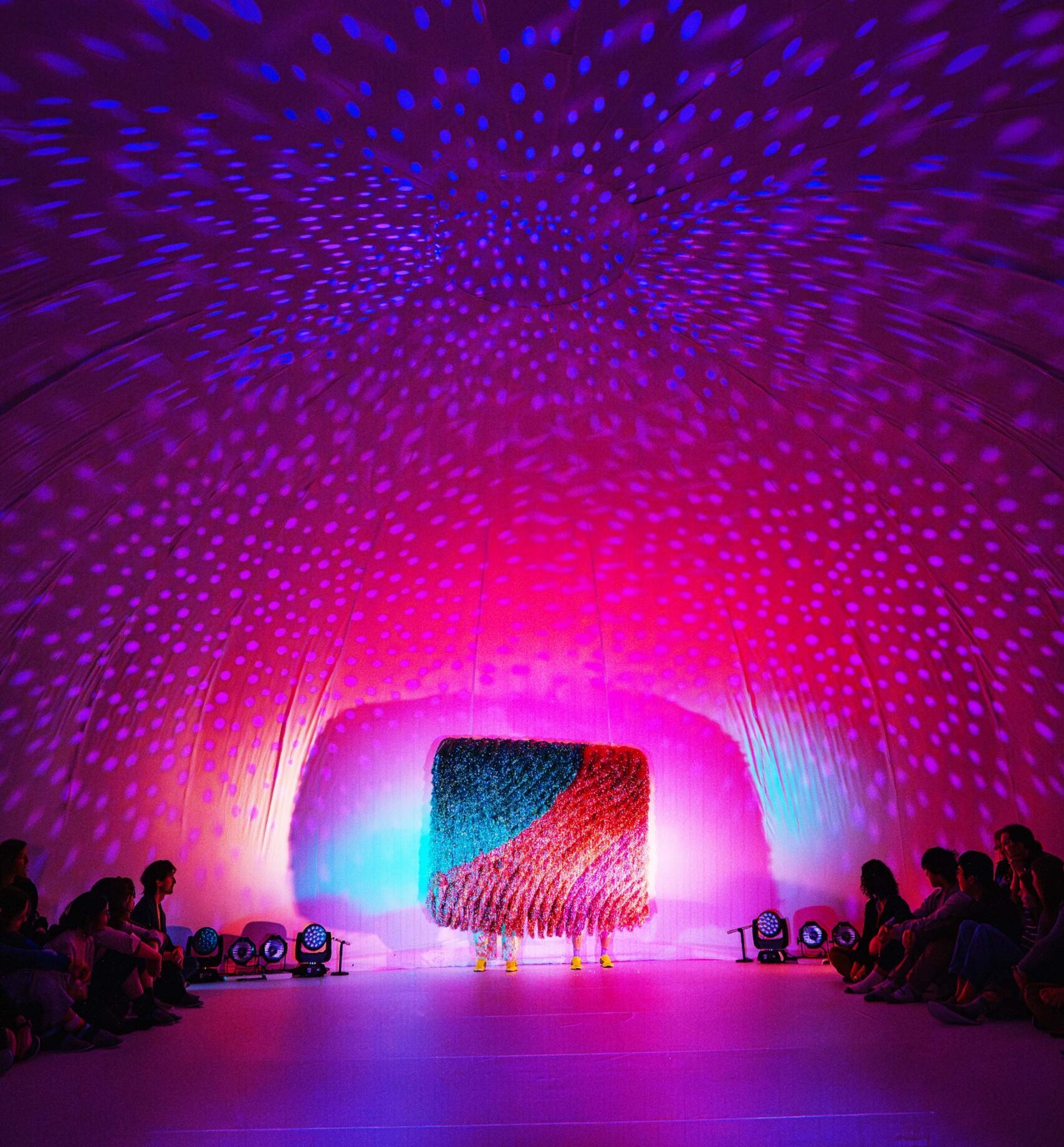 Inside of a large tend and performance space with colored lights reflecting on its ceiling.